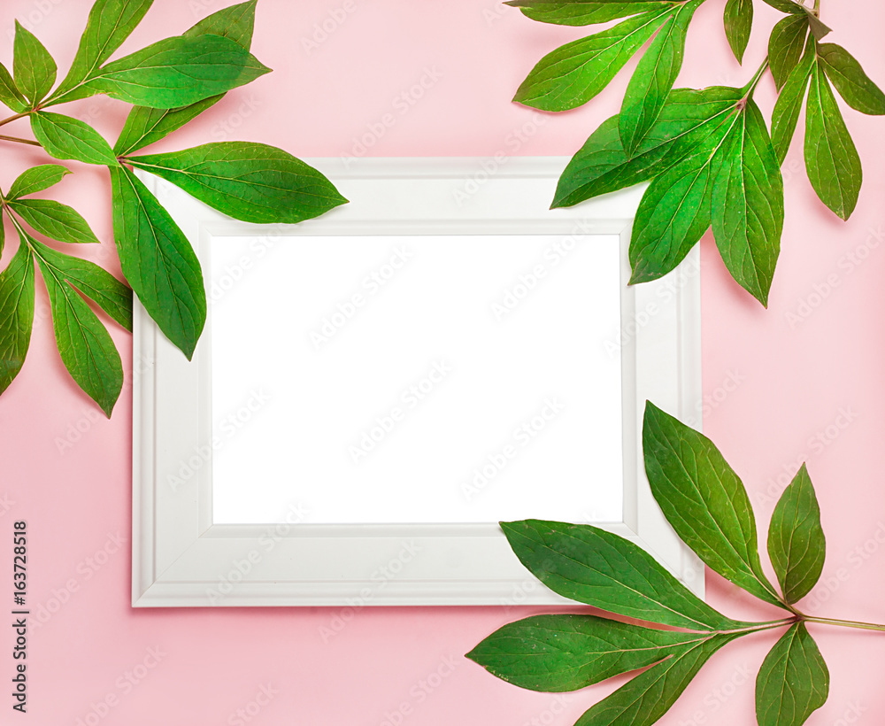 Fototapeta premium white wooden frame on pink background decorated with green leaves, blank space for a text. Top view, flat lay