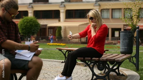Pretty, blonde girl sitting on the bench in the city and doing selfies on smartphone
