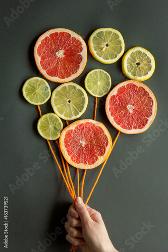 A female hand holds air balloons with orange threads made of citrus slices (lemon, lime, orange, grapefruit) on a black background. Food art concept.