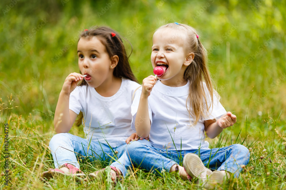 Two little girls relaxing and eating a lollipop with a friend at a park Two little girls relaxing and eating a lollipop with a friend at a park