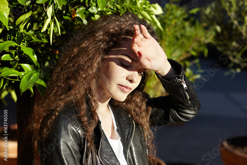 Picture of gorgeous young brunette woman dressed in trendy leather jacket covering face with palm, trying to hind herself from bright sunshine while having rest outdoors in park on summer day