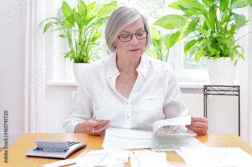 Deutsche Seniorin füllt die Formulare für die jährliche Steuererklärung aus, zuhause an einem Tisch mit Taschenrechner und vielen Rechnungen und Quittungen. photo