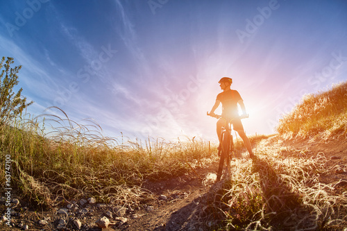 Young cyclist with mountain bicyclist on the path of the field in the countryside against sunrise.