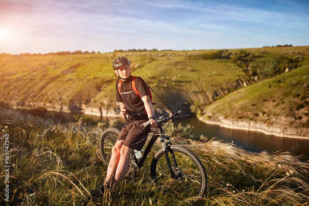 Obraz premium Portrait of the young cyclist standing on the hill above the river against blue sky with clouds.