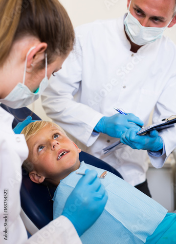 Stomatologist with female assistant are diagnosticating to young patient which is sitting