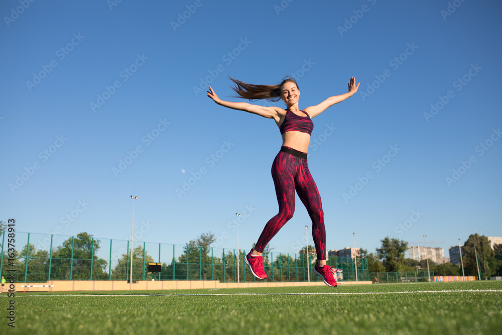 Young happy sportswoman in sportswear jumping and flying on stadium green grass outdoors. She enjoying summer. Healthy lifestyle concept, sport activity.