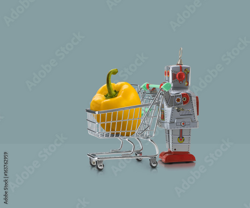 Toy robot pushing miniature shopping trolley with yellow pepper against blue background photo