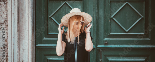 Outdoor close up portrait of model woman in fashionable black dress,and stylish round sunglasses and yellow hat.City lifestyle.Female fashion concept.Retro and vintage filter and colors effect used.