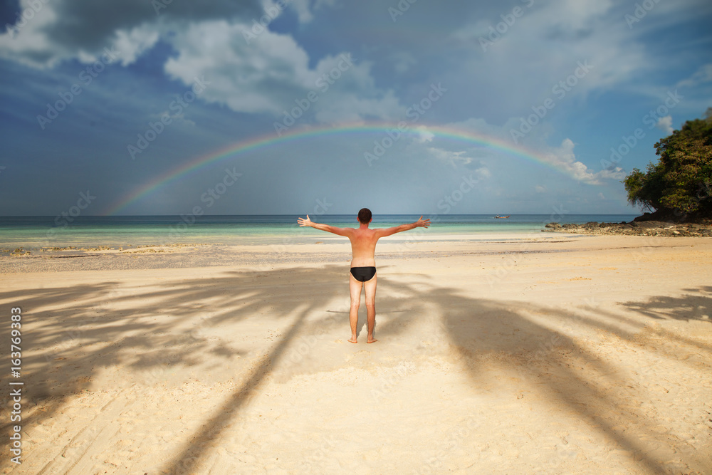 A man stands on Loh Moo Dee Beach, against the background of the sea and the rainbow. Thailand, Phi Phi island.