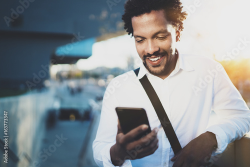 Smiling American African man using smartphone to text message friends at sunny street.Concept of happy young handsome people enjoying gadgets outdoors.Blurred background.
