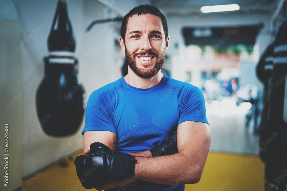 Muscular smiling boxer in gloves standing with arms crossed near punching bag in fitness gym on blurred background.Young caucasian boxer in black gloves.Horizontal.