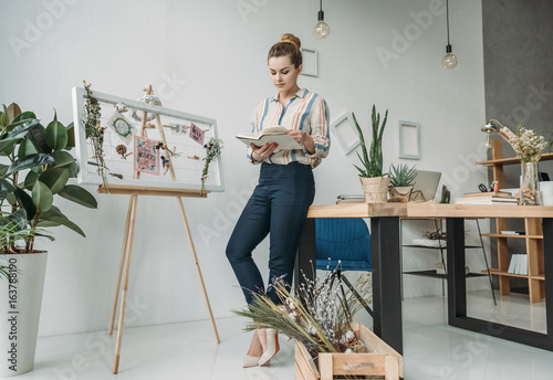 confident attractive female florist holding book and standing at board in office