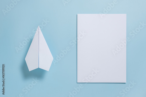 Flat lay of white paper plane and blank paper on pastel blue color background