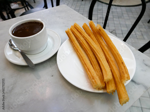 Churros or Spanish fried-dough pastry on white plate served with a cup of hot chocolate as dipping sauce.