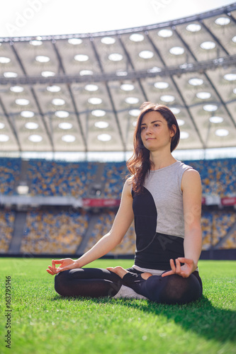 Young happy sporty girl sitting in yoga pose at the stadium. Yoga woman meditating on green grass against the sun