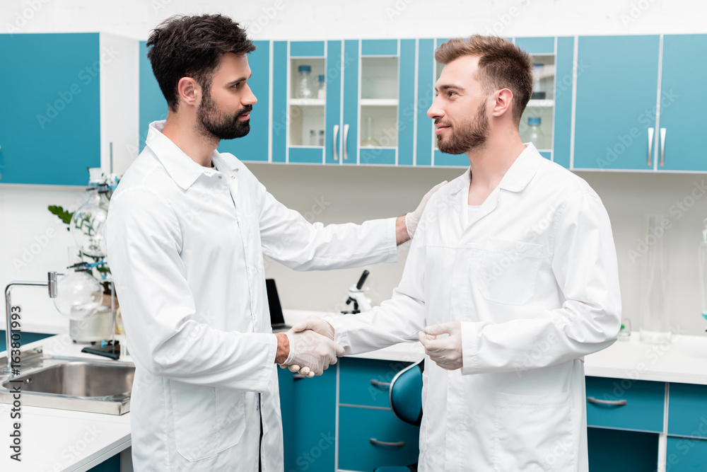 Obraz premium Professional young chemists in white coats shaking hands in scientific laboratory