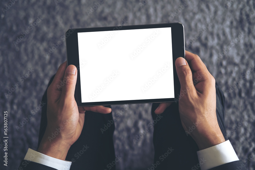 Mockup image of a business man sitting and holding black tablet pc with blank white screen with gray rug in background Mockup image of a business man sitting and holding black tablet pc with blank white screen with gray rug in background