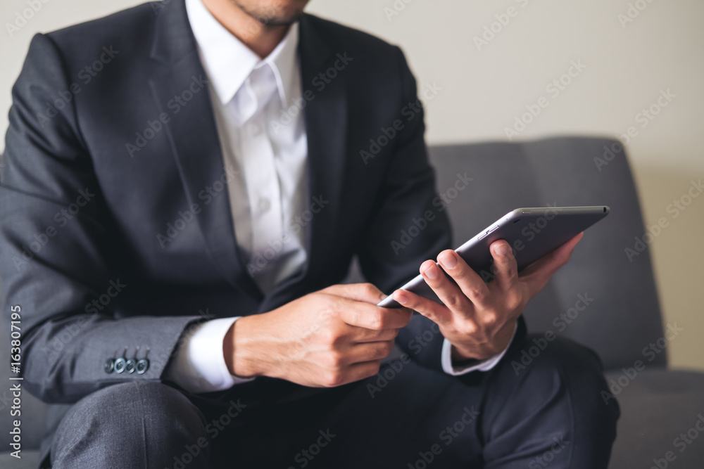 A business man in a gray suit sitting on sofa holding , using and looking at tablet pc for work in office A business man in a gray suit sitting on sofa holding , using and looking at tablet pc for work in office