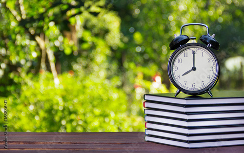 A stack of textbooks and a clock on a wooden table. A stack of books and an alarm clock on a green blurred background. Educational concept. School and lessons.
