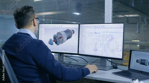  Chief Male Engineer Designs on 3D Model of a Turbine or Engine Part. Second Screen Shows Technical Blueprints. Out of the Office Window Big Factory is Seen.Shot on RED EPIC-W 8K Helium Cinema Camera. photo