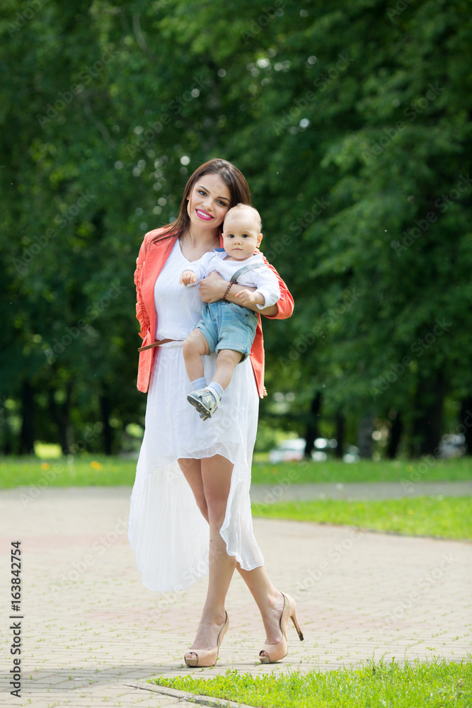 A little boy with dark eyes in a white shirt and denim shorts in the hands of his beautiful happy mother in a green park in the summer A little boy with dark eyes in a white shirt and denim shorts in the hands of his beautiful happy mother in a green park in the summer