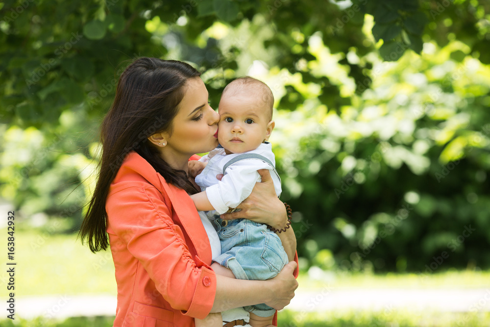 A little boy with dark eyes in a white shirt and denim shorts in the hands of his beautiful happy mother in a green park in the summer