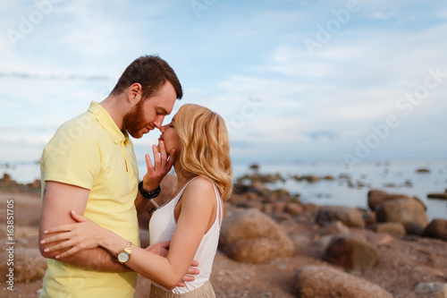 Young beautiful smiling couple hugging on the shore amid rocks and sea, enjoy the sunset and freedom. Male to prepare for a kiss. Journey to the sea.