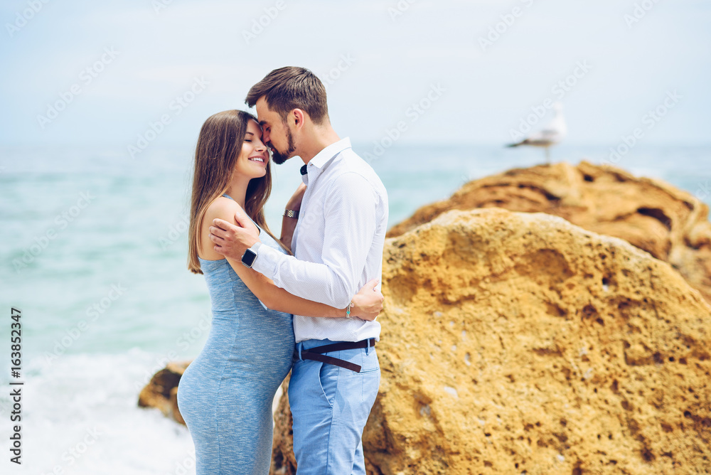 A beautiful pregnant woman in a blue dress and her courageous and handsome husband in a shirt and blue trousers leaned to each other against the background of the rock and the sea.