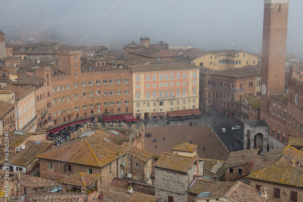 Torre del Mangia in Piazza del Campo and tupical ref roofs of Siena in the thick fog. Tuscany, Italy.