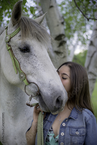 Vertical photo of dappled white horse wearing green bridle with girl kissing side of face. Best friends with birch tree background. Young brown haired girl with eyes closed wearing denim jean jacket. 