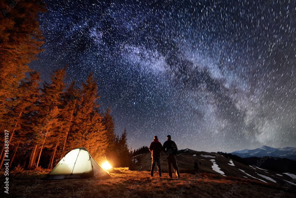 Two male tourists have a rest in the camping near the forest at night. Guys standing near campfire and tent under beautiful night sky full of stars and milky way, enjoying night scene. Long exposure