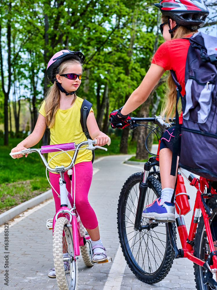Obraz premium Bicycle path and sign with children. Girls wearing bicycle helmet with rucksack ciclyng ride. Kids are on white bike lane. Alternative to urban transport. Oncoming traffic. Unexpected meeting of two