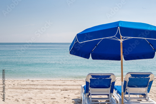 Two blue beach lounge chairs under a blue umbrella facing the ocean on a clear blue day