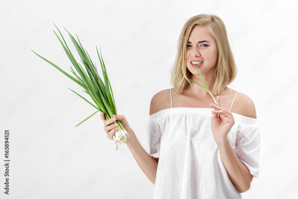 Naklejka premium Beautiful blond woman in a white blouse eating green bitter onion on white background. Health and vitamins