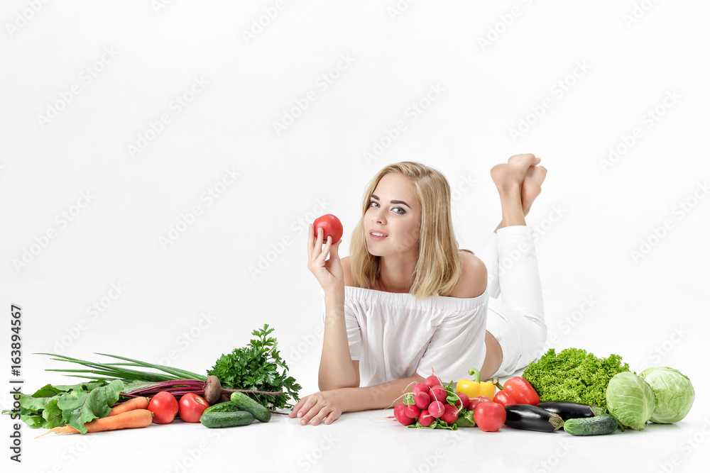 Beautiful blond woman in white clothes and lots of fresh vegetables on white background. Girl holding tomato Beautiful blond woman in white clothes and lots of fresh vegetables on white background. Girl holding tomato