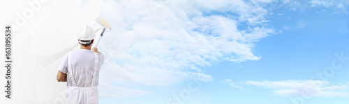 Rear view of painter man painting the blue sky on blank wall, with paint roller isolated on white big space panorama