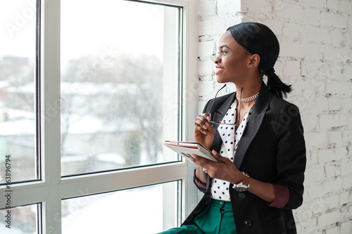 Smiled girl with dark shiny skin sitting on the windowsill, holding the tablet and looking out the window.