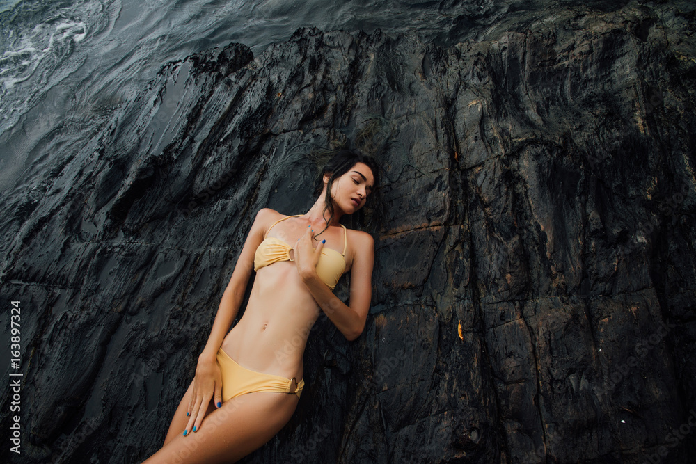Sexy brunette in bikini lies on a large rock on ocean coast