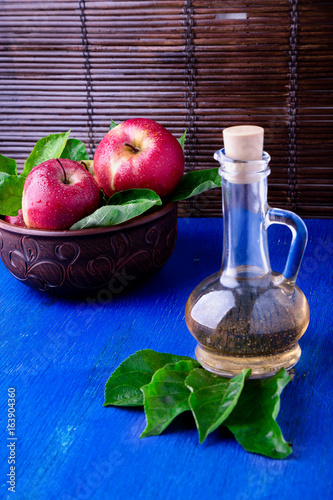 Apple cider vinegar in glass bottle on blue background. Red apples in brown bowl.