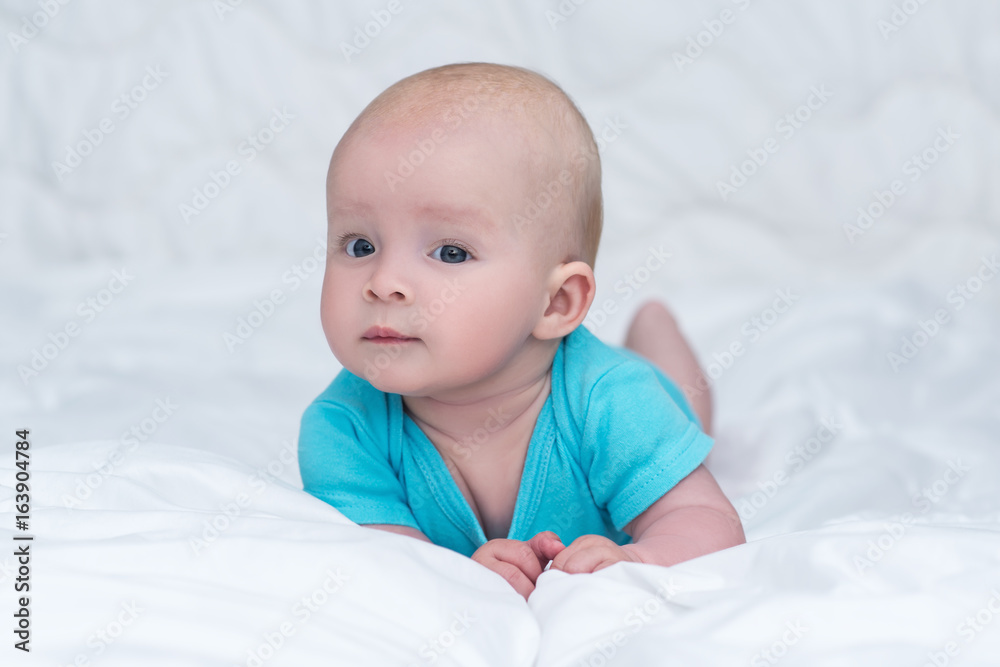 adorable baby girl or boy in blue shirt with big blue eyes lying on her stomach on soft blanket and looks into a camera with surprised, indoors
