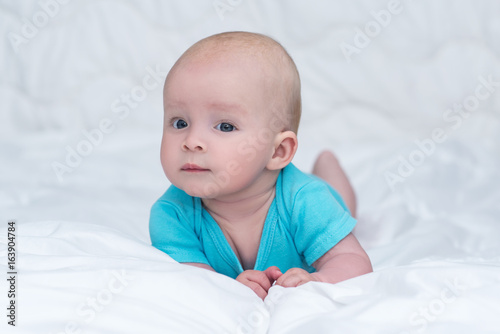 adorable baby girl or boy in blue shirt with big blue eyes lying on her stomach on soft blanket and looks into a camera with surprised, indoors