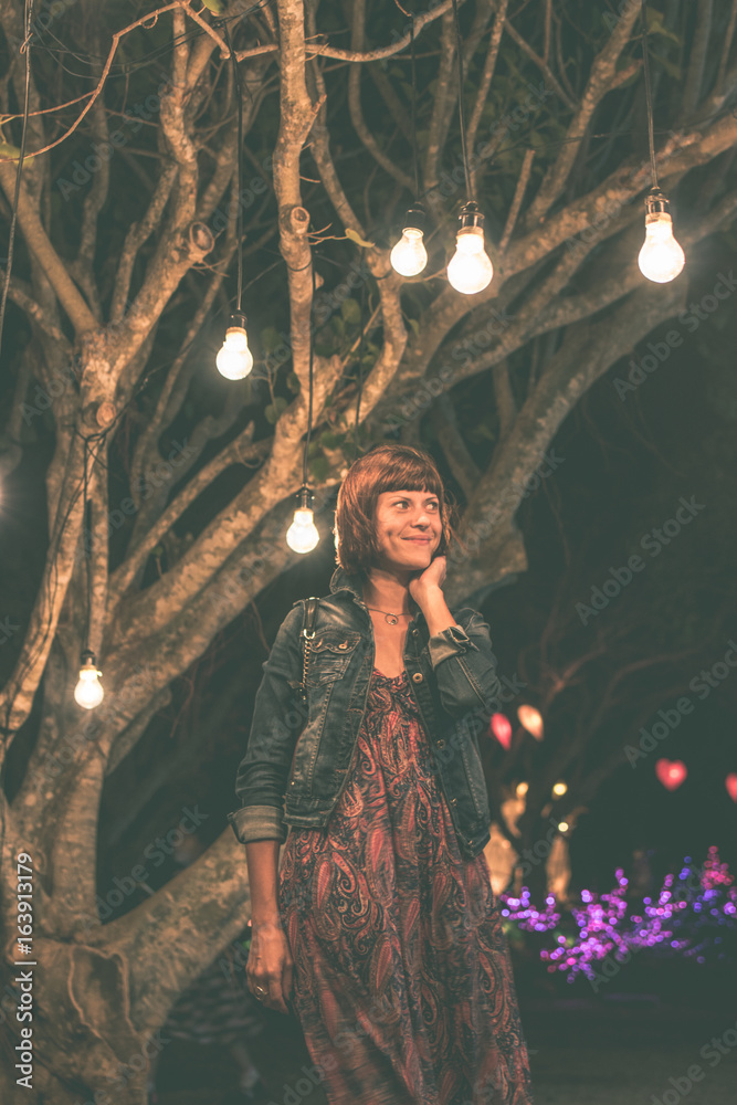 Woman among decorative outdoor string lights hanging on tree in the park at night time. Bali island, Indonesia.