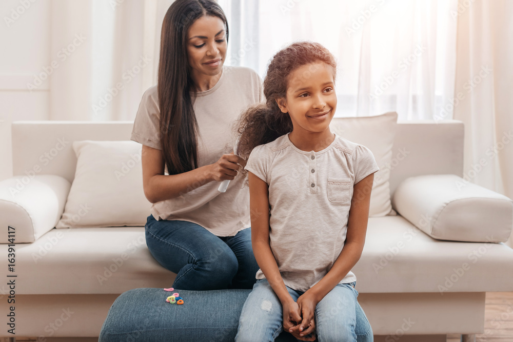 Smiling afro american mother combing hair to little daughter at home