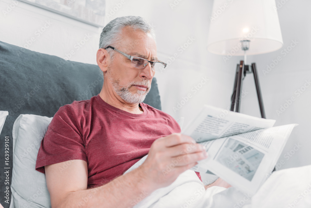 Obraz premium focused senior man reading newspaper while lying in bed at home