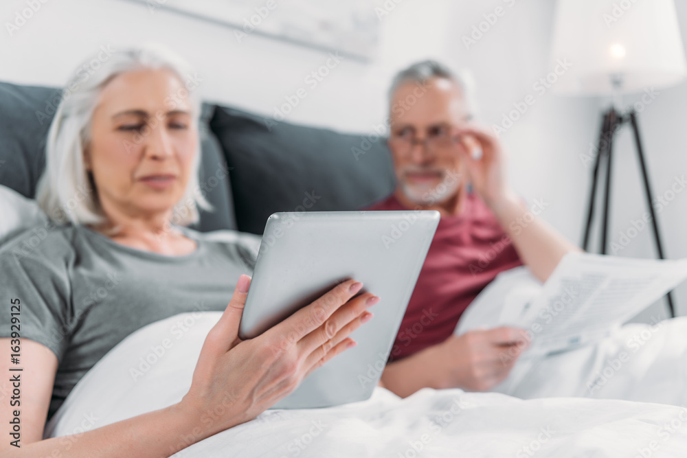 selective focus of woman using tablet while husband reading newspaper in bed selective focus of woman using tablet while husband reading newspaper in bed