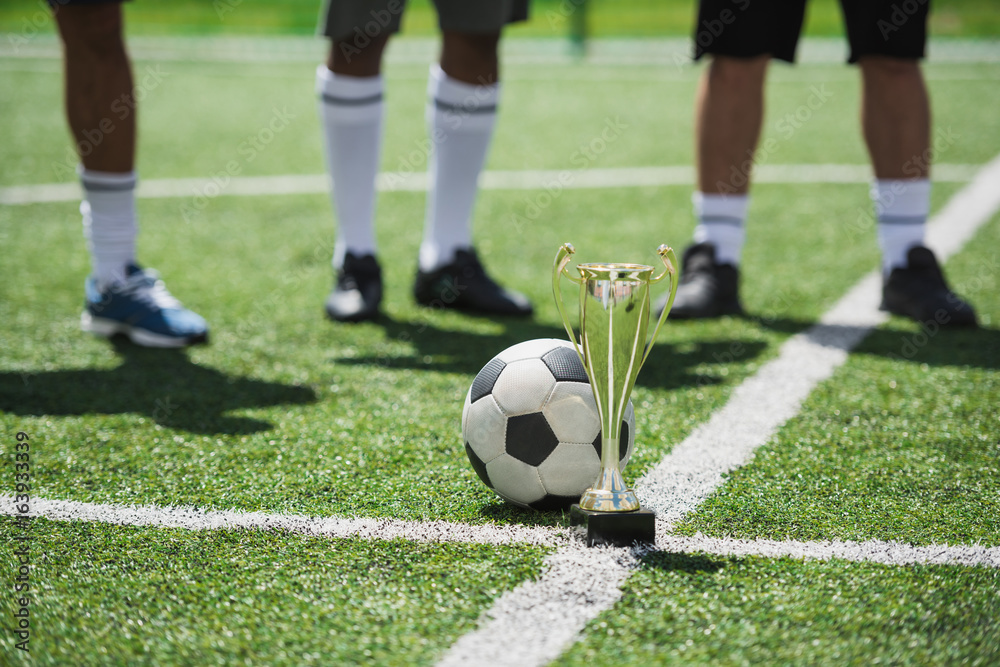 goblet and soccer ball on football field with players standing behind