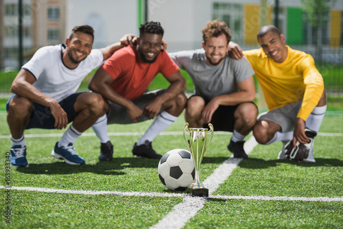 smiling multiethnic soccer team with goblet on soccer pitch after game