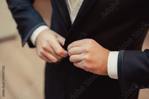 Elegant groom in wedding jacket wearing white shirt and turquoise tie. Groom's hands on blue suit.