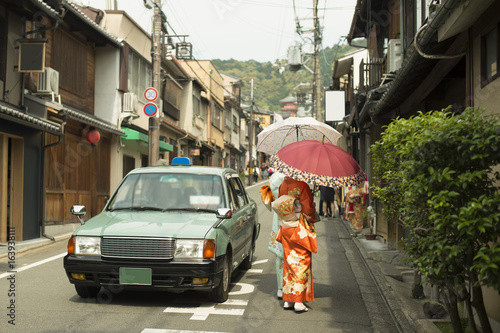 Vintage street scene in Kyoto province, Japan with taxi car and females wearing Yukata and handling traditional umbrellas.