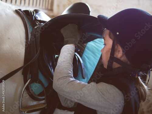 The girl tightens the girth on the horse. A girl in a helmet saddles a horse.